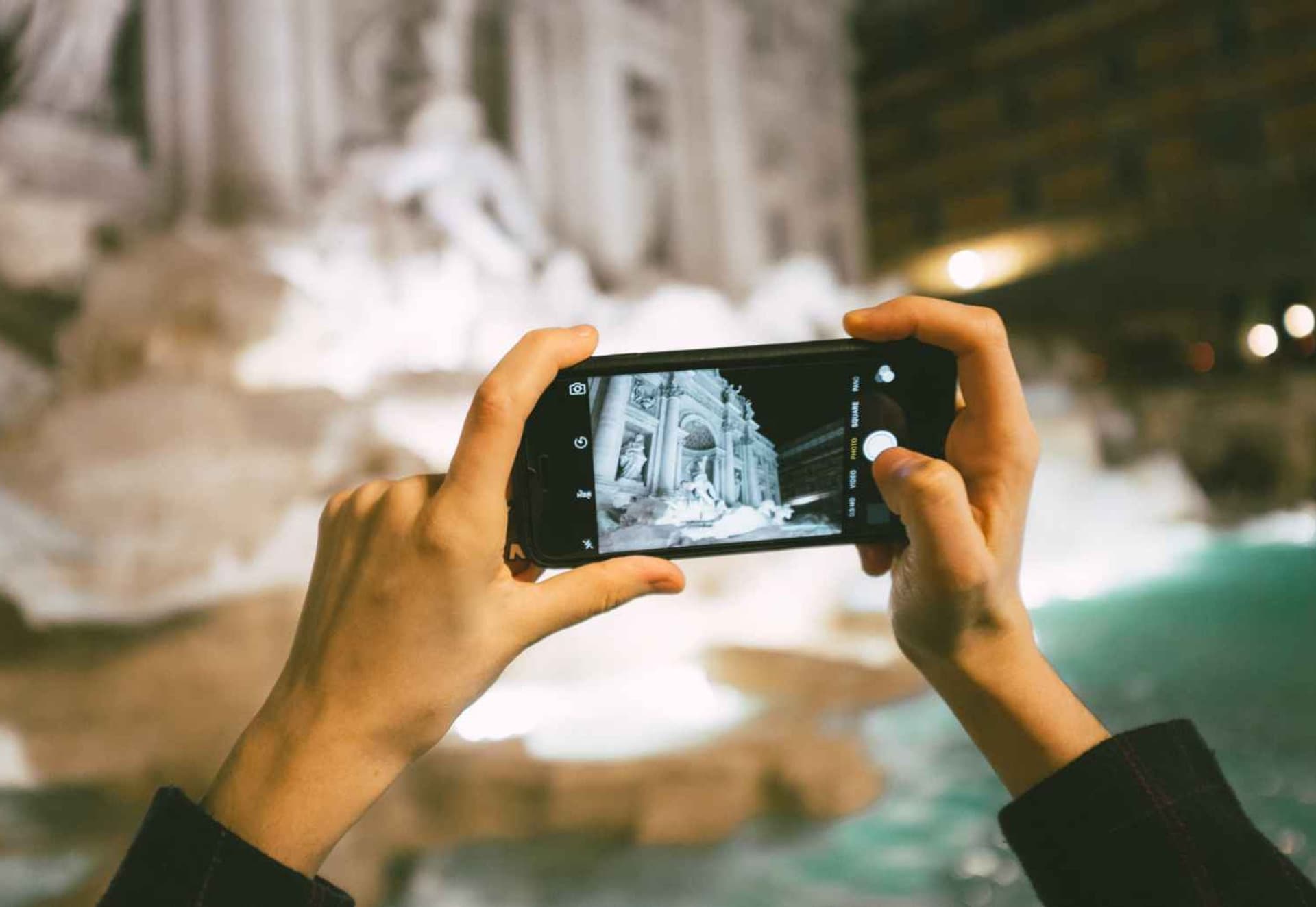 A person capturing a photograph of the iconic Trevi Fountain in Rome, surrounded by tourists and beautiful architecture.