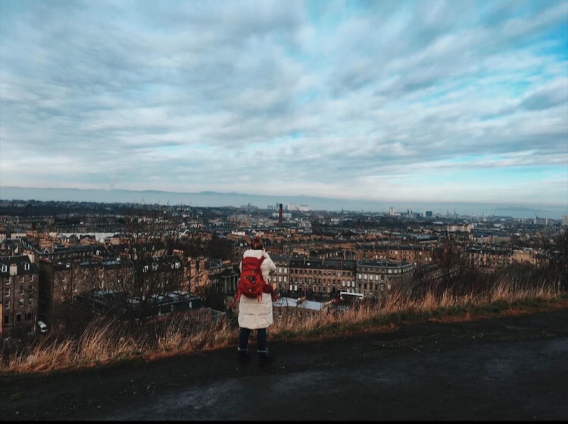 A person stands on a hill, gazing at a sprawling cityscape below, with buildings and streets visible in the distance.